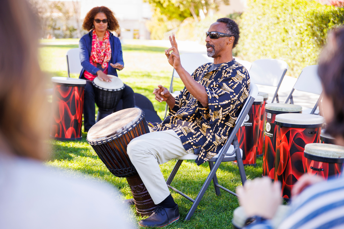 A Black man holding a drum speaks to participants of a drum circle at Dexter Lawn.