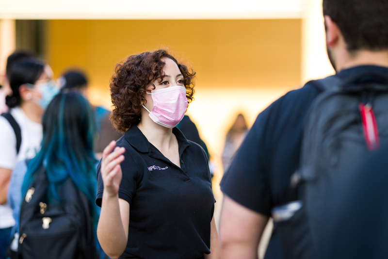 A person in a mask and black polo shirt speaks to a fellow student 