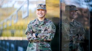 A man in military camoflage smiles in front of a building window. 