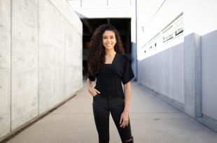 A woman with curly dark hair wearing an all-black outfit stands in a concrete outdoor hallway.