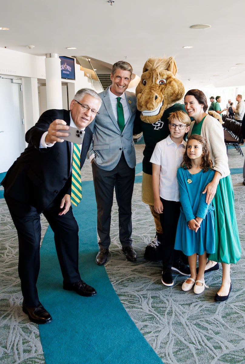 Cal Poly president Jeffrey Armstrong holds a phone while posing with mascot Musty, athletic director Carter Henderson, his wife and two kids.