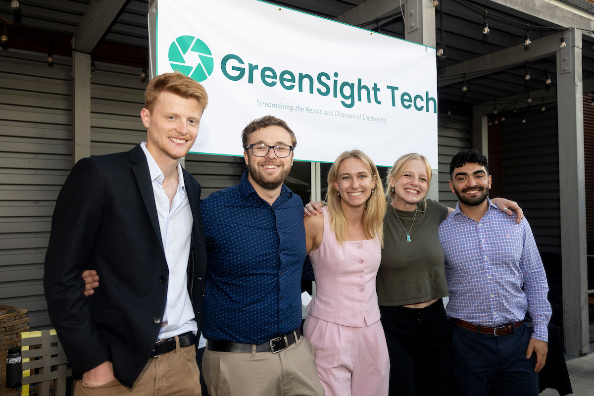 Five members of the GreenSight Tech team smile and stand together in front of a poster for the company.