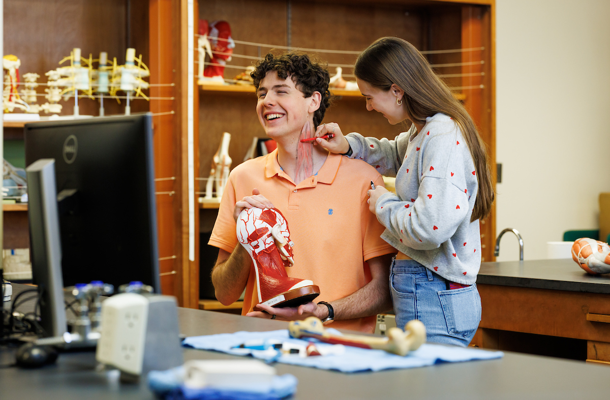 A student smiles while another student paints anatomically correct muscles on his neck.