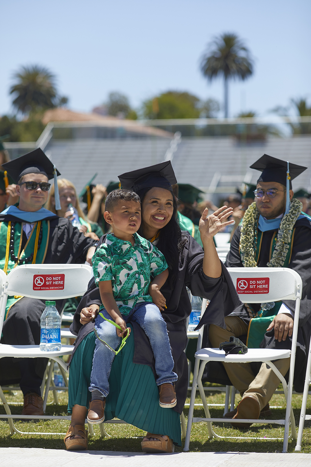 A graduating student smiles as she sits with fellow graduates. Her young child is on her lap.