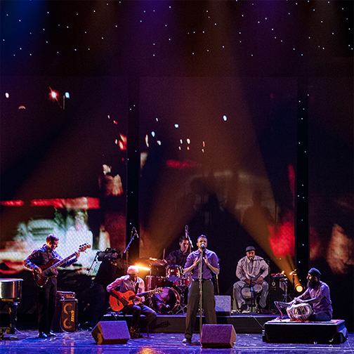 Aditya Prakash is pictured on stage with blue and red lights behind him as his band stands behind him.