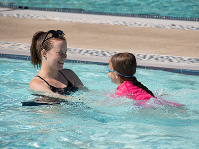 An adult and child in a pool, with the adult holding the child's hands and smiling as the child swims toward her.