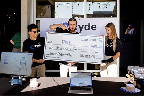 Three students standing behind a booth holding an oversize check for their startup idea, Ryde 