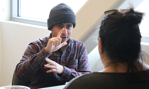 Mechanical engineering student Yosef Towfique, seated with a flannel shirt and gray beanie, talking using ASL to another person.