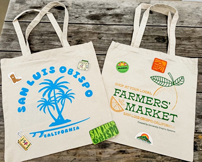 Two tote bag examples. One says San Luis Obispo with blue illustrated palm trees and the other says Farmer's Market with different designs in green and gold.