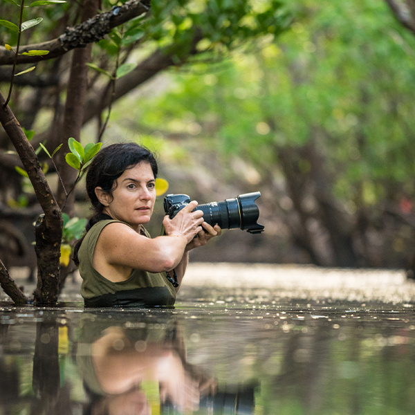 Woman holding a camera waist-deep in water in a river.