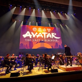 Orchestra plays on stage with Avatar on screen behind.