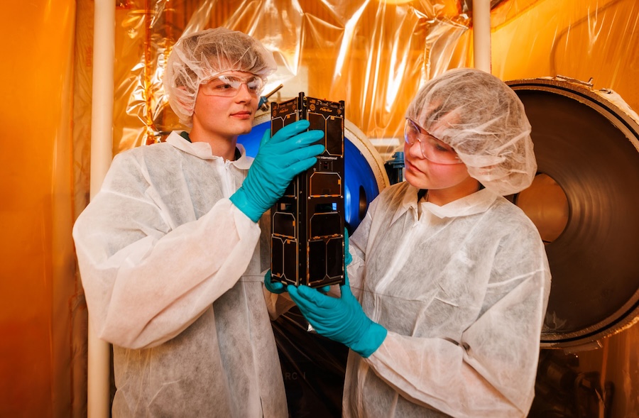 Two engineering students wearing protective glasses and lab coats hold a CubeSat within a lab environment.