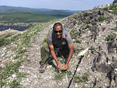 Nishanta Rajkaruna on the top of a rocky mountain kneeled with flowers in hand.