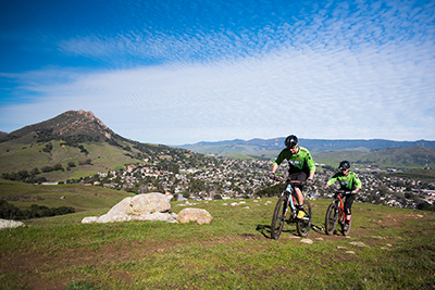 Two people ride mountain bikes up a hill with campus in the background