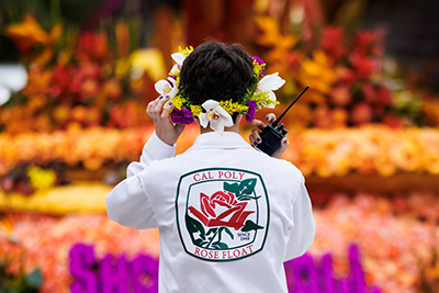A student wearing white coveralls with a Cal Poly Rose Float wears a flower crown and holds a radio near a Rose Parade float covered in colorful flowers