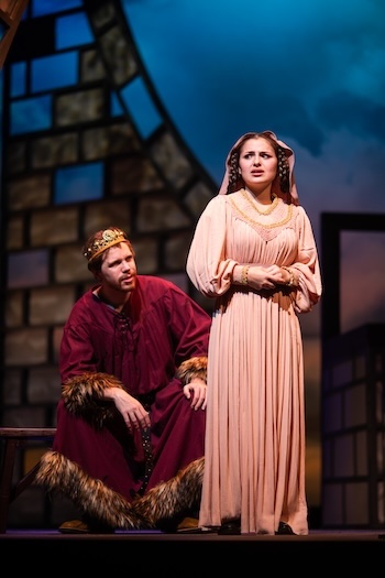 A man in a robe with fur detailing and a crown sits on stage with a woman standing facing away.