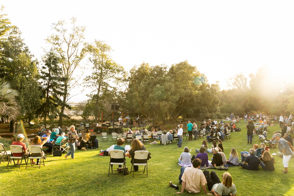 Dozens of people sit on chairs and blankets on a lush green lawn facing a stage with tall trees surrounding it