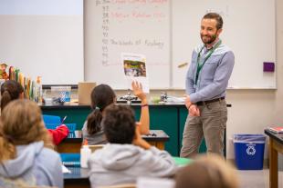 A young teacher smiles as students raise their hands in a classroom