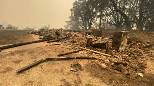 The rubble from a fire-damaged building, with charred trees and a smoky sky in the background