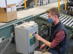 A student wearing a blue face mask kneels in front of a gray metal box and pushes a green button.