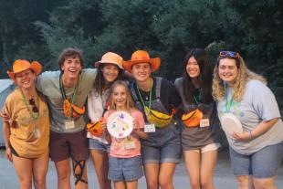 Six college students in colorful hats and fanny packs pose for a photo with a smiling child at an outdoor campsite.