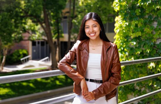 Christina Benitez Ruiz leans on a railing and smiles on a sunny day outside on campus.