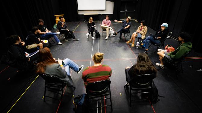 A group of students and a faculty member sit in chairs in a circle in a black box theater