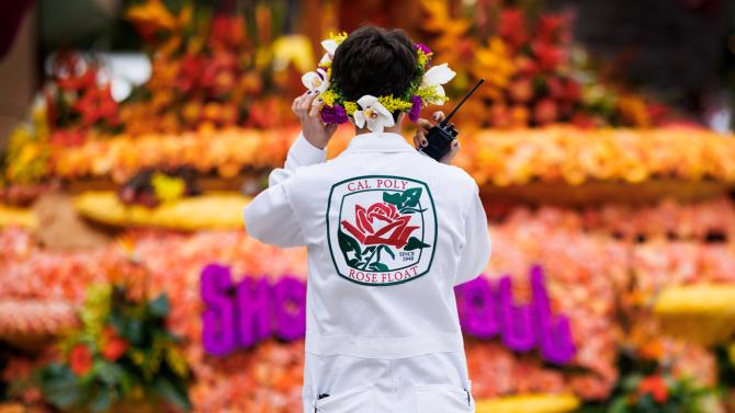 A person wearing a white set of Cal Poly Rose Float coveralls and a flower crown holds a radio in front of a colorful Rose Parade float