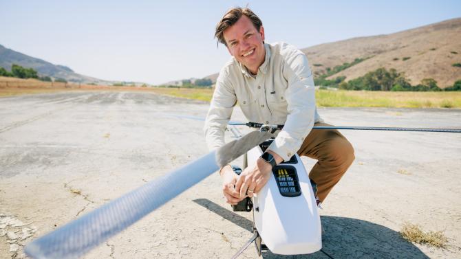 A student smiles while kneeling next to a drone on a runway