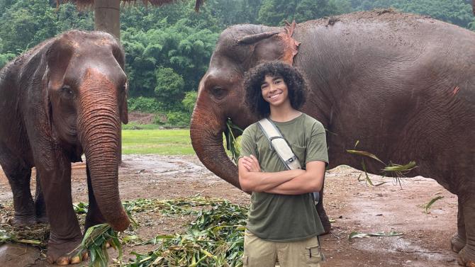 A student smiles in front of two Asian elephants