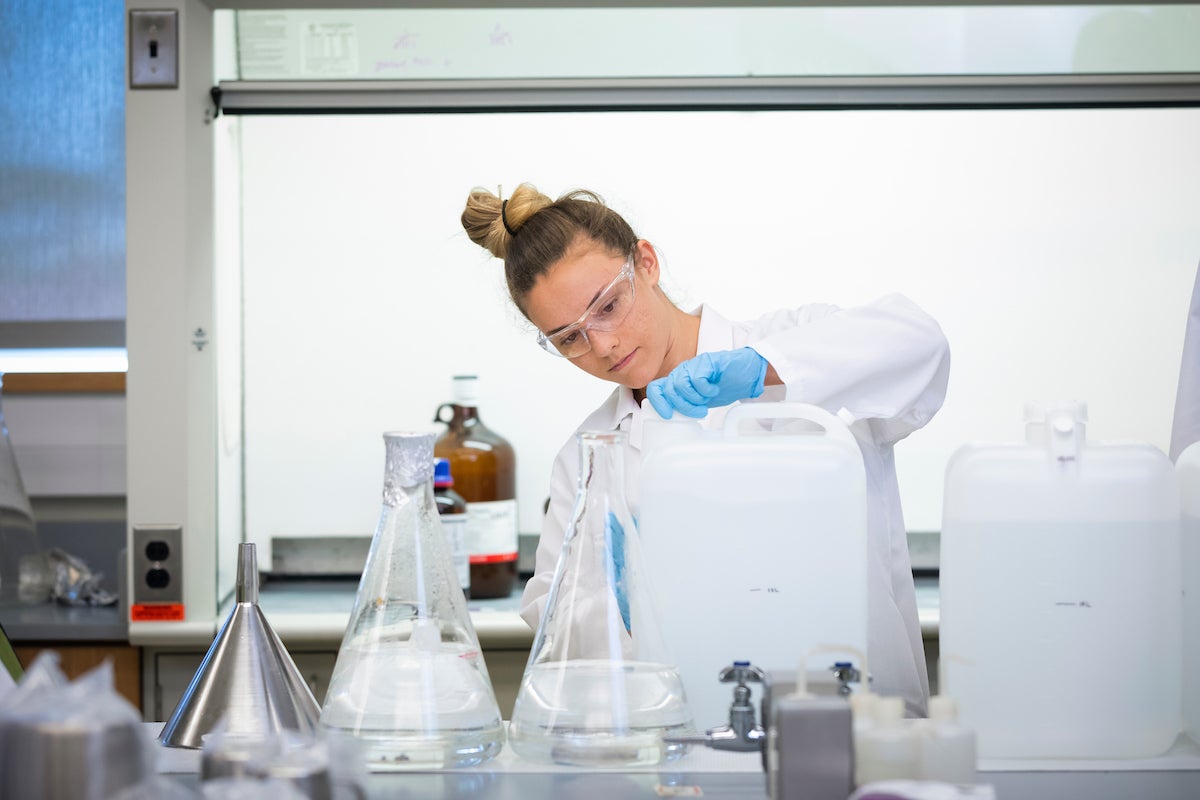 A woman in a white lab coat pours hand sanitizer into a container. 