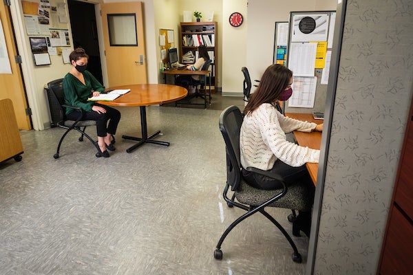 Two students wearing masks sit inside a room. One student is reading a binder at a small table while the other student is six feet away and working on a computer.