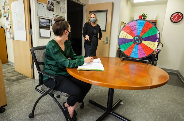 A student in a green blouse and face mask talks to a blond woman in a black cardigan and gray face mask.