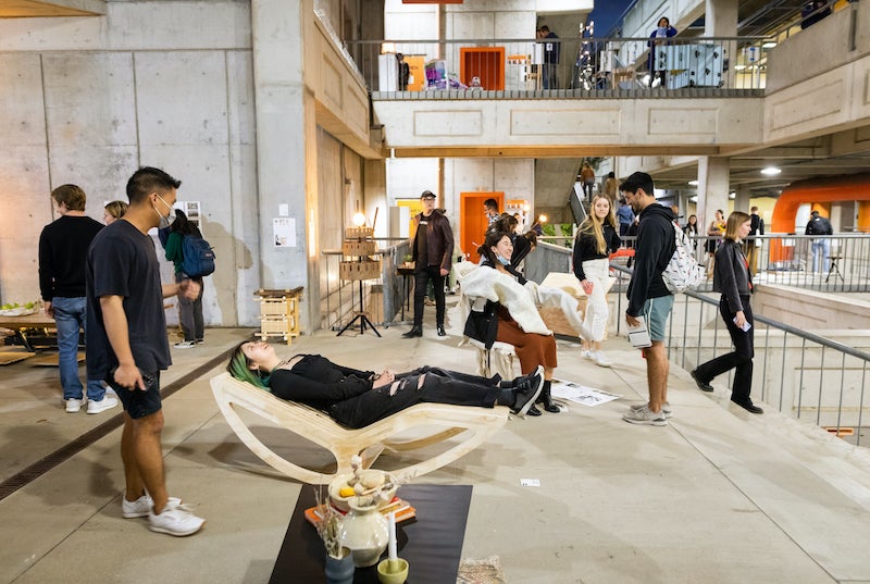 Students and faculty socialize and try out unique furniture. A student in the middle is lying down on a wooden recumbent chair.