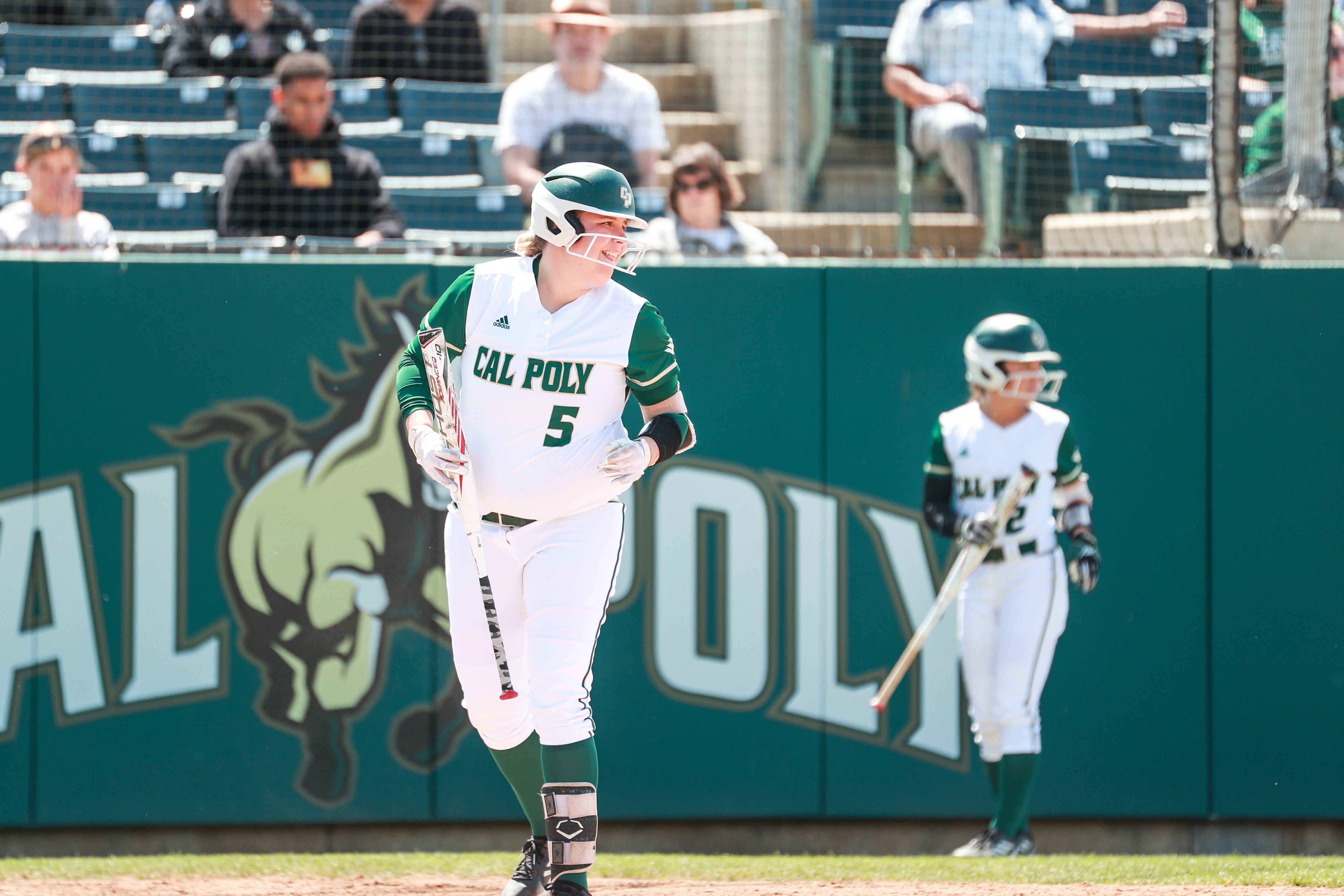 A woman wearing a Cal Poly softball uniform holds a bat and grins as she jogs around the field.