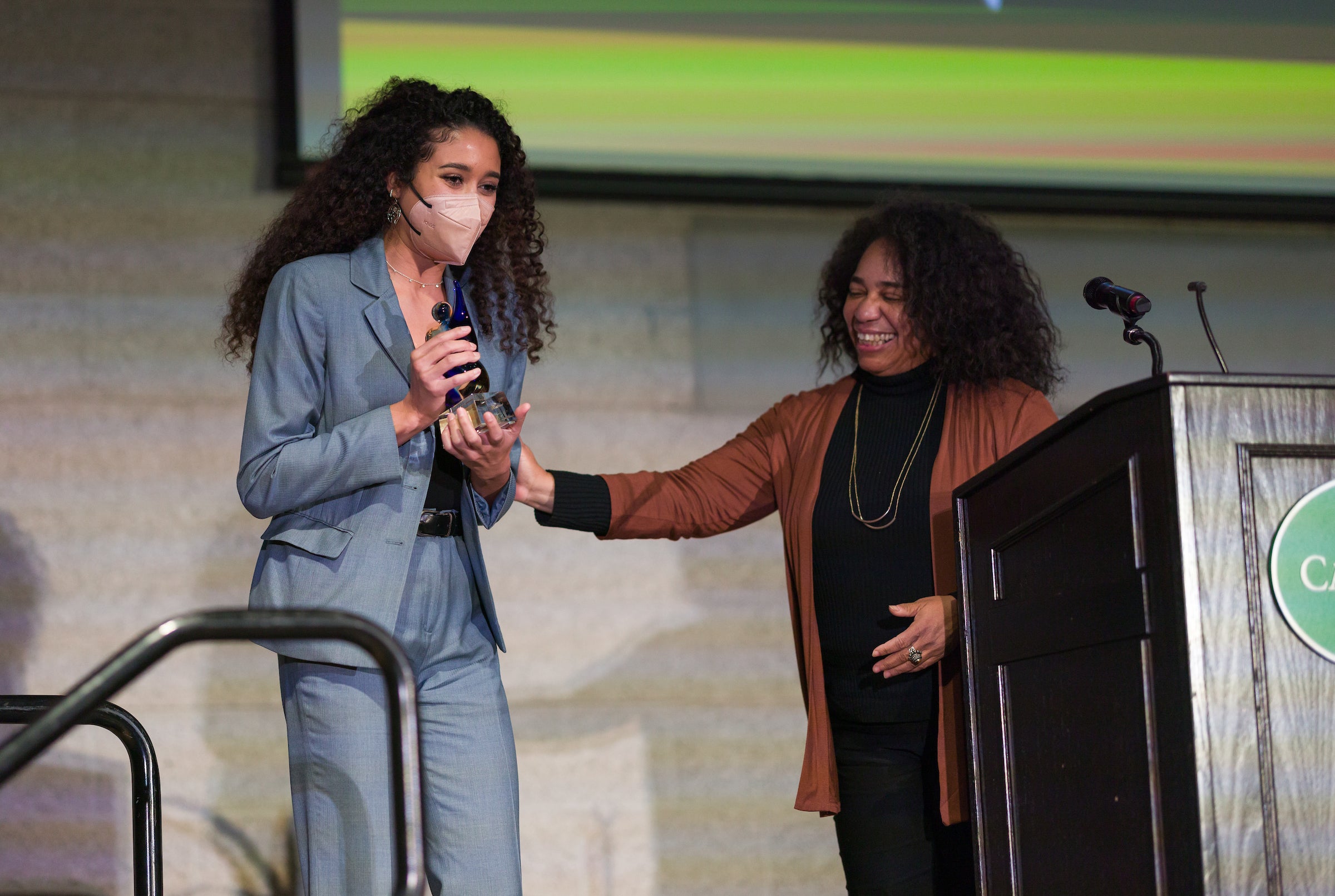 Two women wearing dressy clothes stand on a stage near a podium.