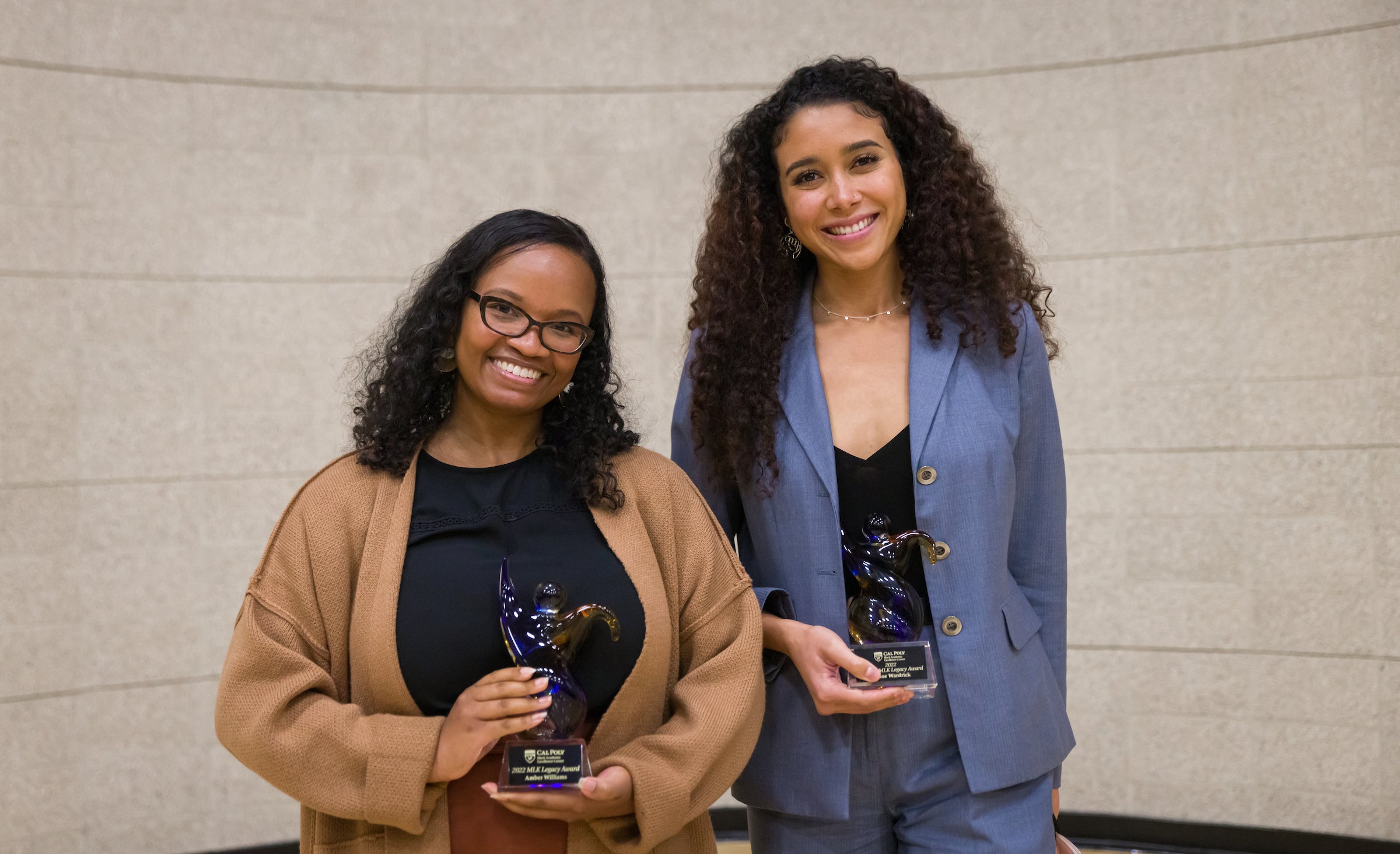 Two women wearing suits and holding awards stand together and smile. 