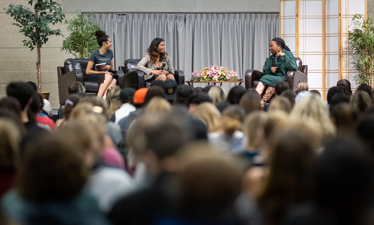 In front of a crowded auditorium, two young women with microphones sit on stage opposite Burke