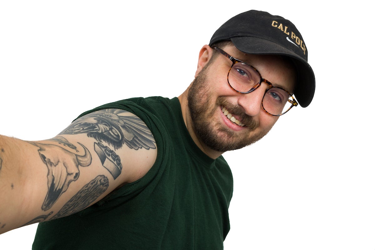 A young man in a cap shows off several arm tattoos, including a prominent Lego block