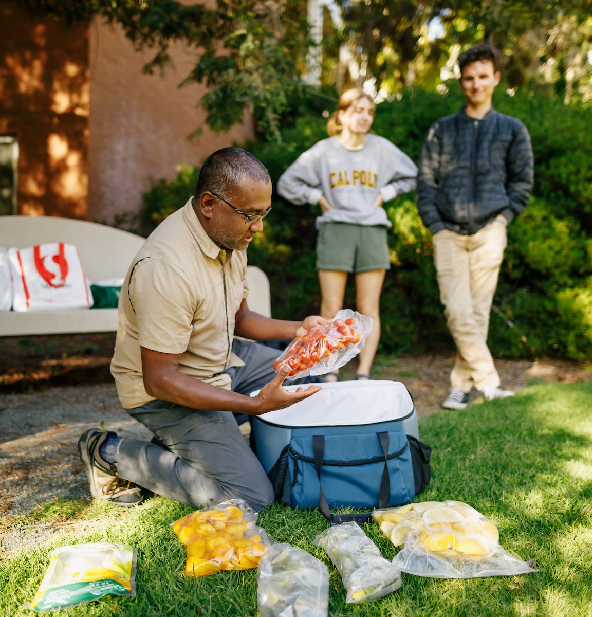 Professor Nishi Rajakaruna kneels on the ground to pull Ziploc bags of fruits out of a cooler. He is surrounded by students.