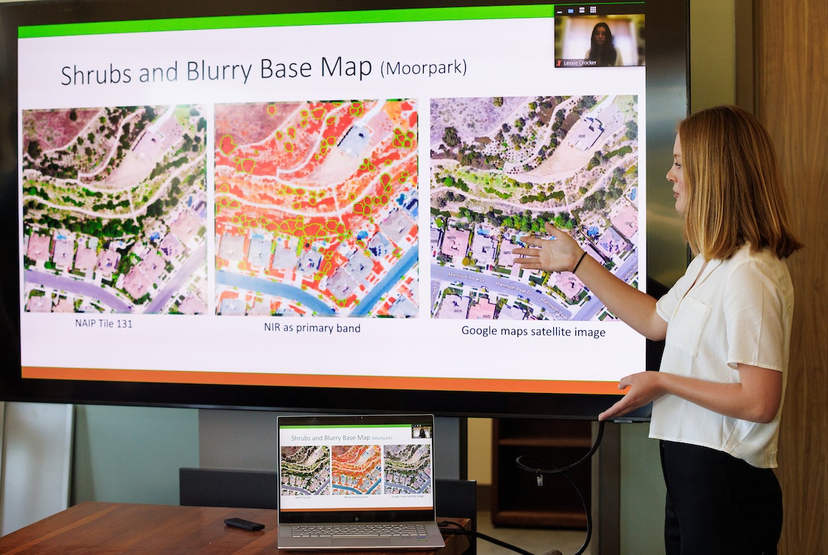 A student in a white shirt speaks and gestures at a screen behind her, which is showing a slide from a PowerPoint presentation.