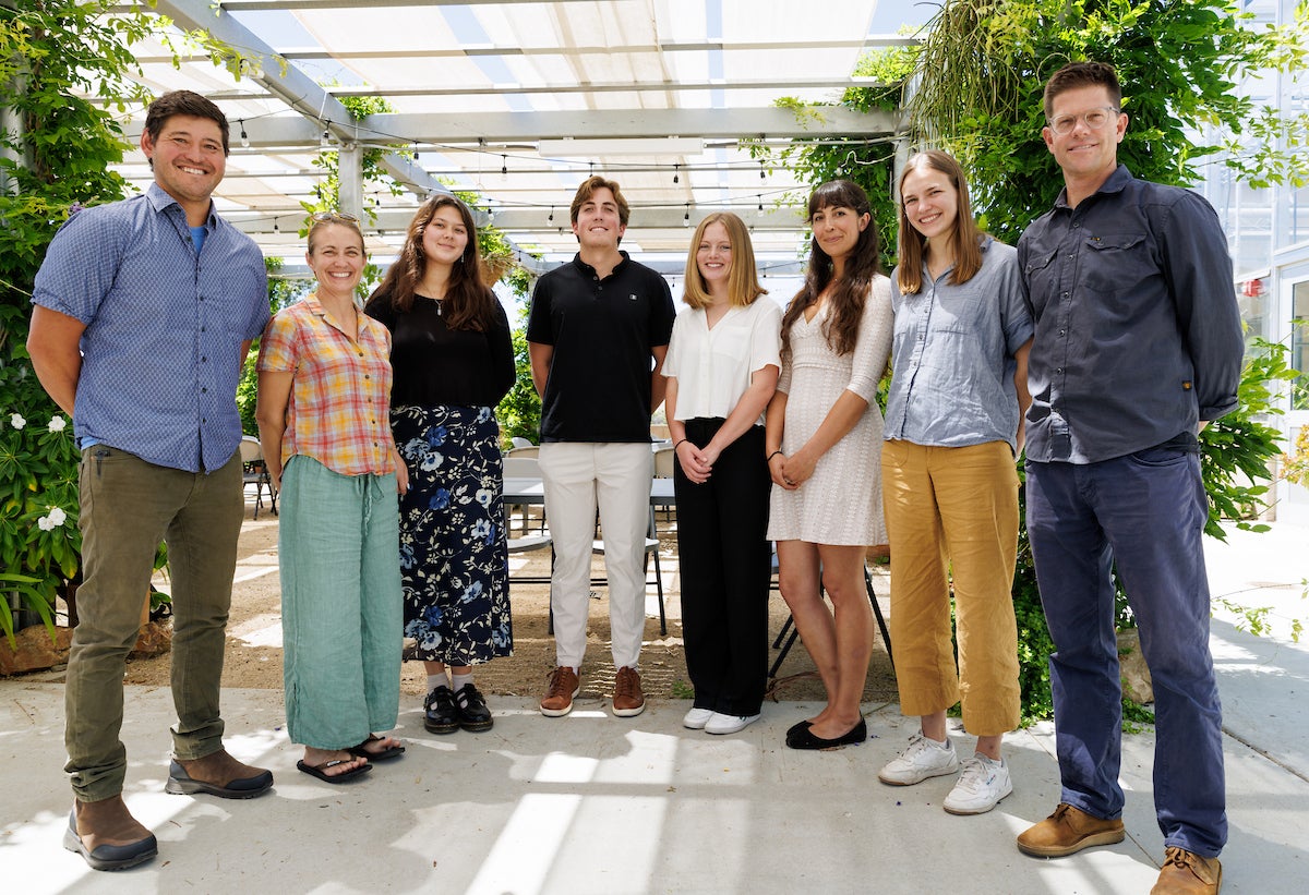 A group of four students and three professors stand in a semicircle and smile for a group photo.