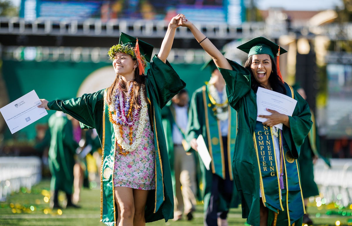 Two students in graduation robes hold hands and smile after they cross the stage.