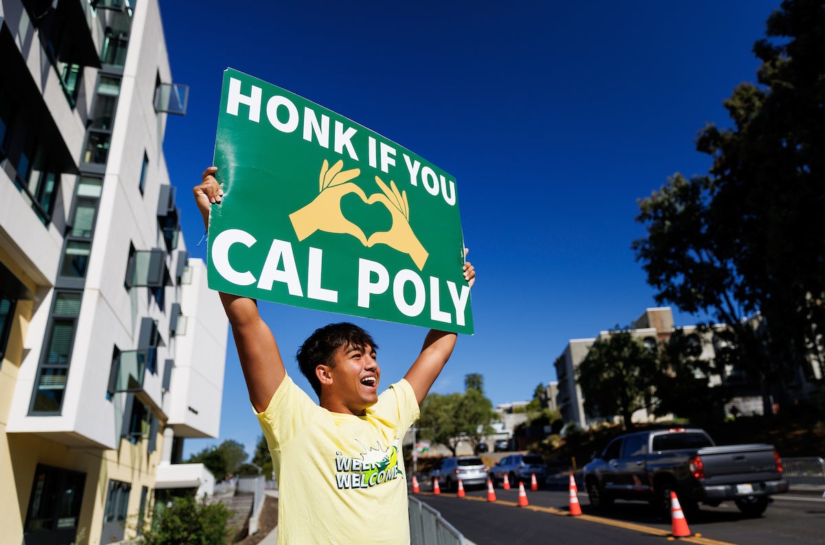 A student smiles and holds a sign that says "Honk if you love Cal Poly" on the side of Grand Avenue.