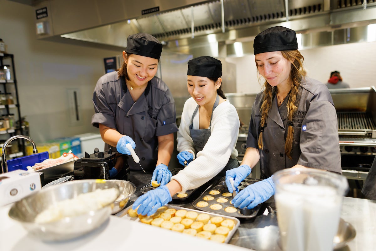 Three students assemble tamal asado in muffin tins in a culinary lab.