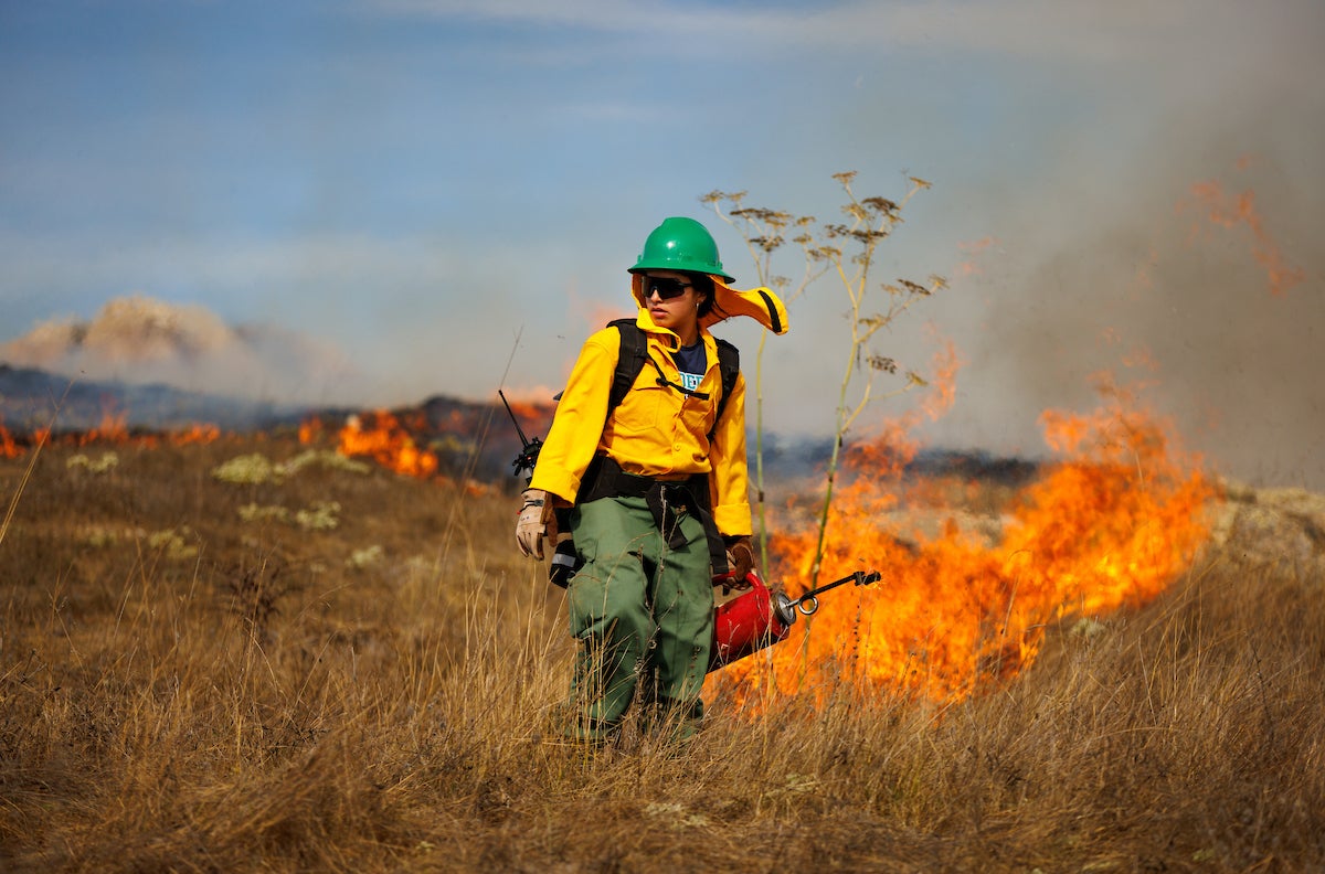 A student wears protective firefighting gear and carries a drip torch, surrounded by flames.