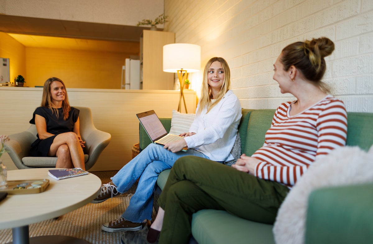 A student sitting on a green couch holds her computer screen to an advisor also sitting on the couch, while a third person sits in a chair and smiles.