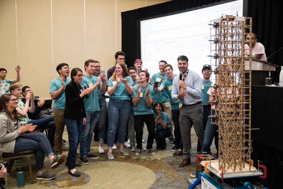 A crowd of students in bright blue shirts claps and a wooden structure stands to the right of the frame.