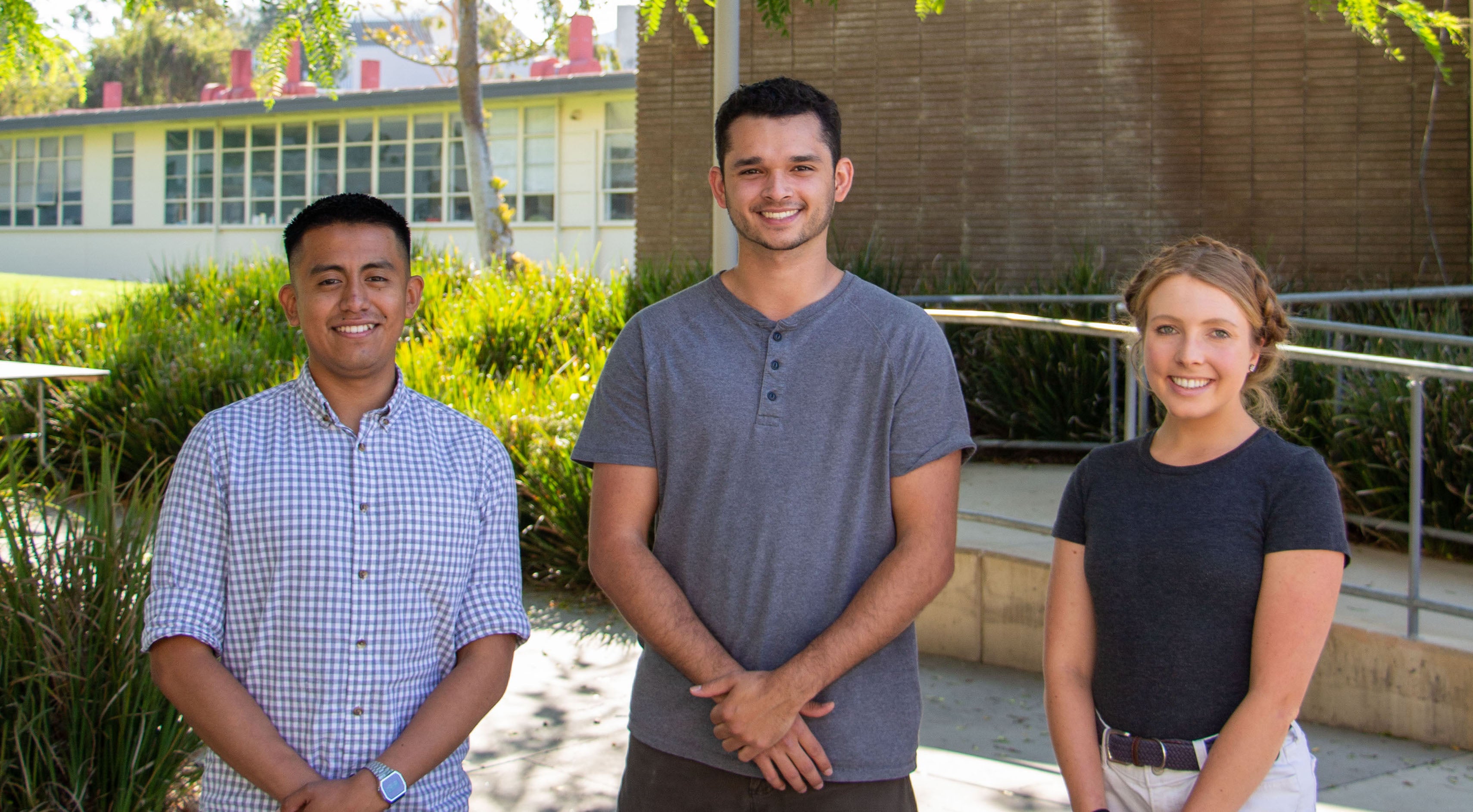 Three students stand with their hands clasped in front of them. The two male students are on the left and center and the female student is on the right.