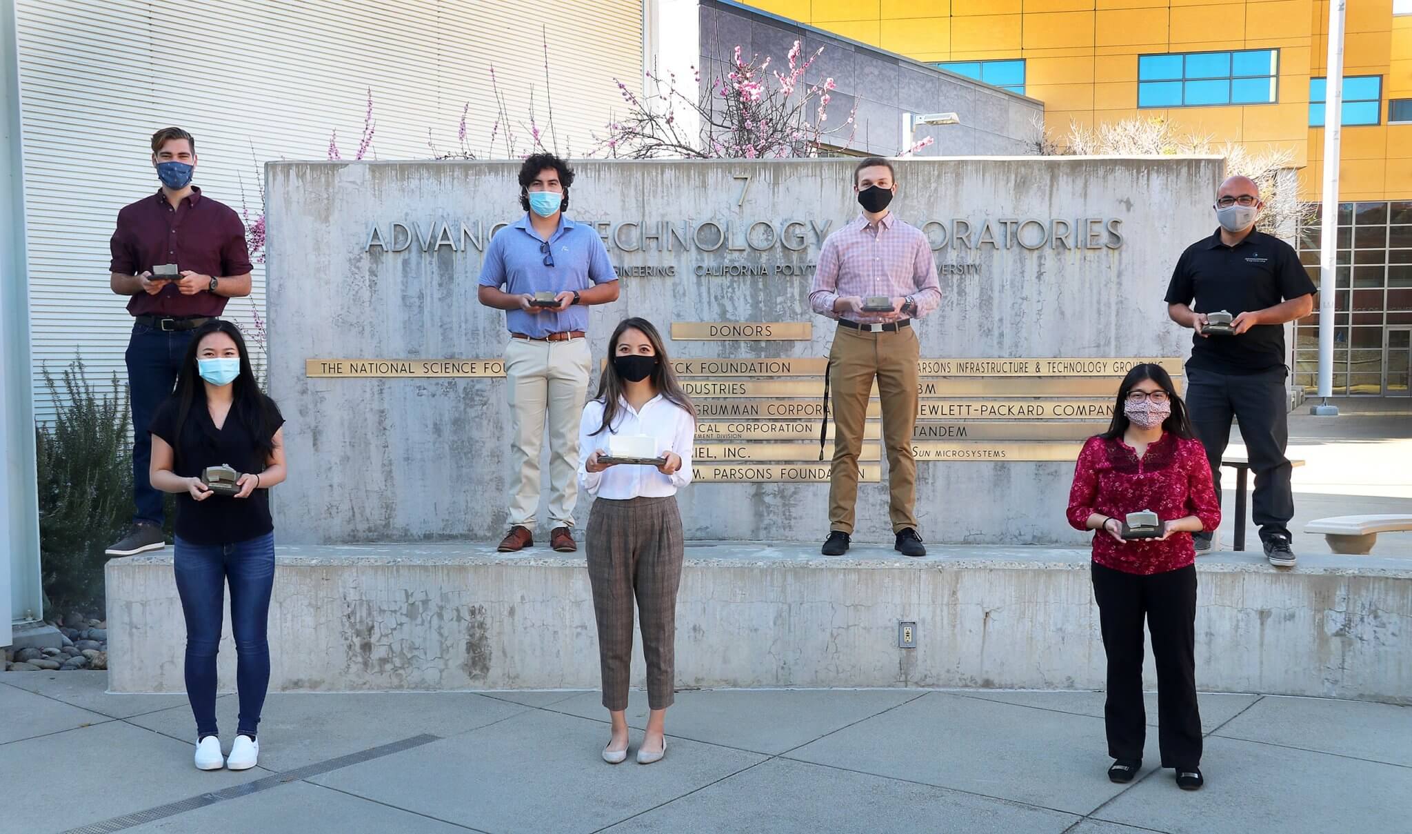 Seven male and female presenting students stand distanced outside on concrete steps, each holding their own small trophy.